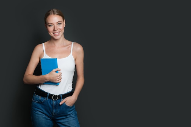 Mujer joven sosteniendo un libro azul y sonriendo sobre un fondo gris