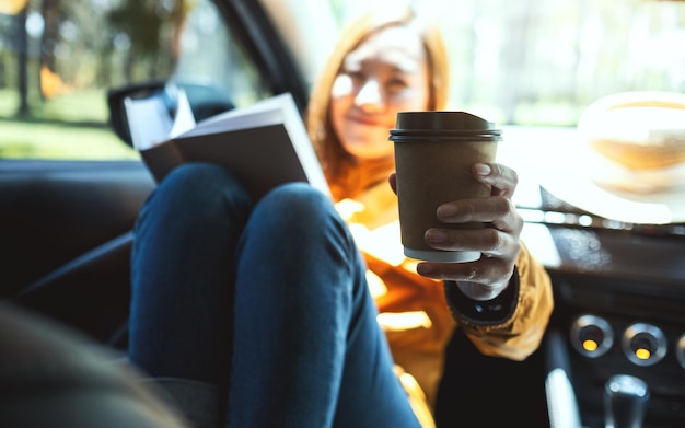 Una mujer joven sosteniendo un libro y dando una taza de café en el auto