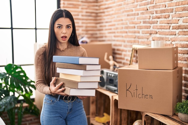 Mujer joven sosteniendo libros en casa con cara de shock que parece escéptica y sarcástica sorprendida con la boca abierta