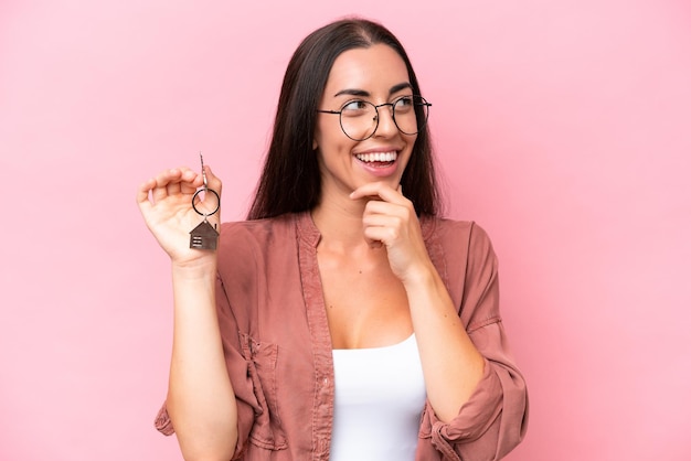 Mujer joven sosteniendo llaves de casa aisladas en un fondo rosa pensando en una idea y mirando de lado