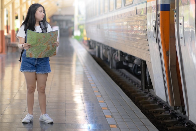 Una mujer joven sosteniendo un mapa de pie en el andén mirando el tren