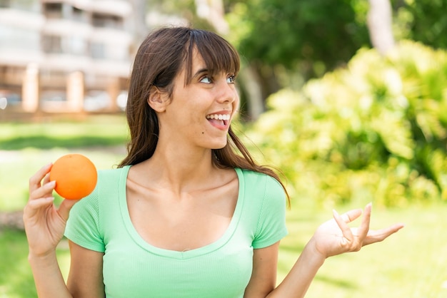 Foto mujer joven sosteniendo una naranja al aire libre con expresión facial sorpresa