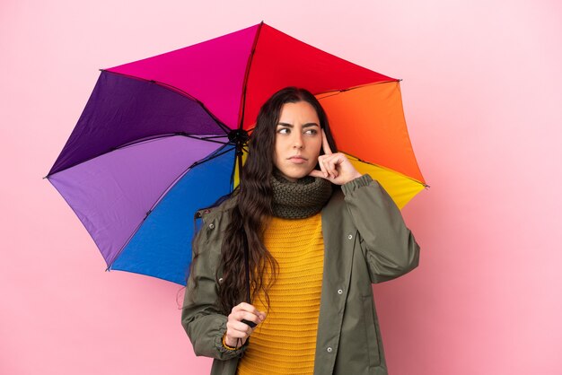 Mujer joven sosteniendo un paraguas aislado sobre fondo rosa pensando en una idea