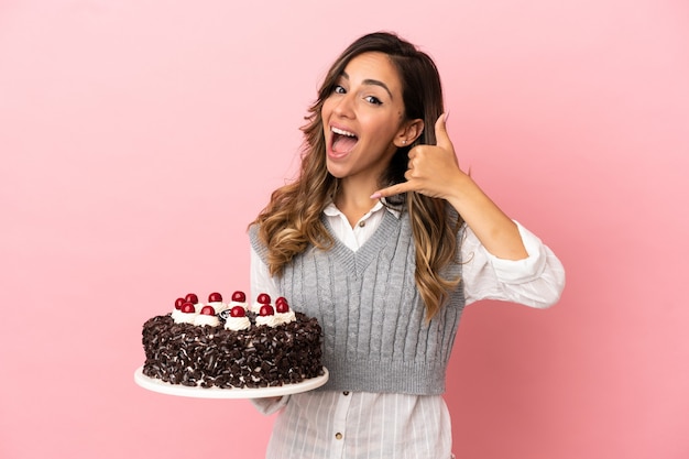 Mujer joven sosteniendo pastel de cumpleaños sobre fondo rosa aislado haciendo gesto de teléfono. Llámame señal
