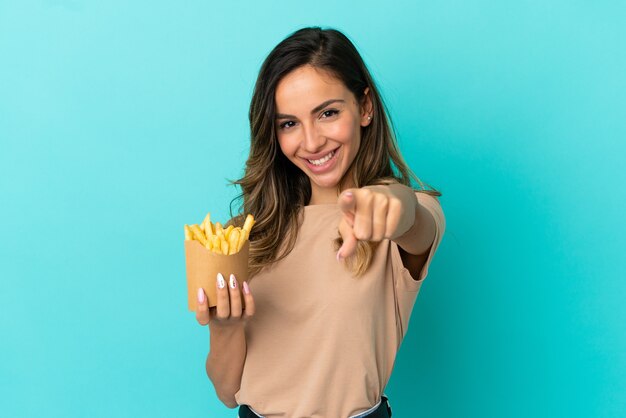 Mujer joven sosteniendo patatas fritas sobre fondo aislado apuntando hacia el frente con expresión feliz