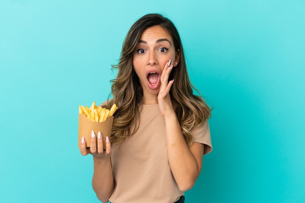 Mujer joven sosteniendo patatas fritas sobre fondo aislado con sorpresa y expresión facial conmocionada