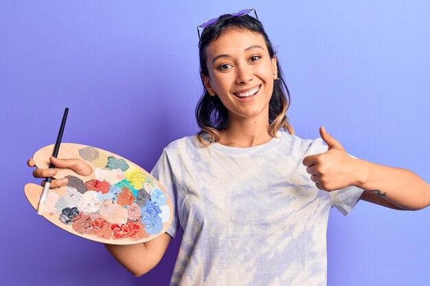 Foto mujer joven sosteniendo pincel y paleta sonriendo feliz y positivo pulgar hacia arriba haciendo excelente y signo de aprobación