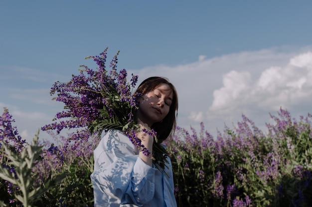 Mujer joven sosteniendo un ramo de flores púrpuras de pie entre un campo de flores silvestres en frente del cielo