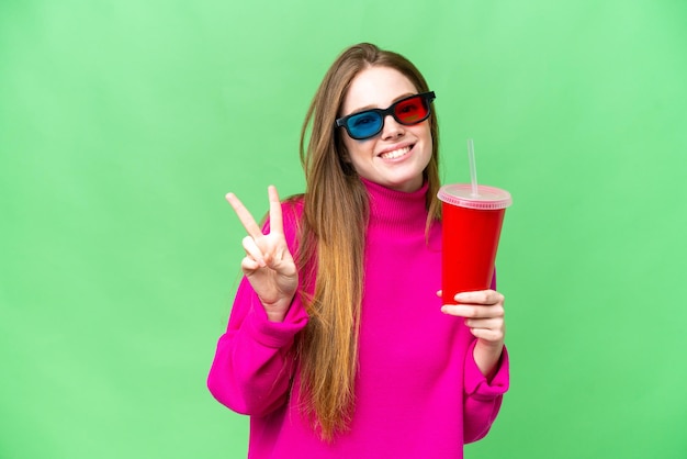 Mujer joven sosteniendo refresco mientras ve una película en 3D sonriendo y mostrando el signo de la victoria