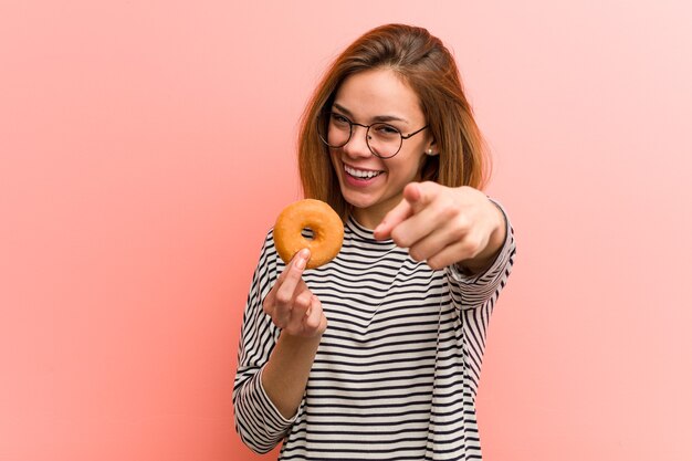 Mujer joven sosteniendo una rosquilla alegre sonríe apuntando al frente.