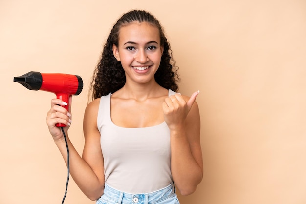 Mujer joven sosteniendo un secador de pelo aislado en un fondo beige apuntando hacia un lado para presentar un producto