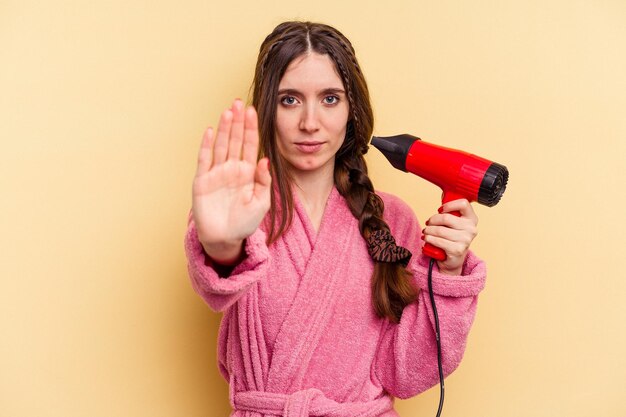 Mujer joven sosteniendo un secador de pelo aislado sobre fondo amarillo de pie con la mano extendida mostrando la señal de stop que te impide