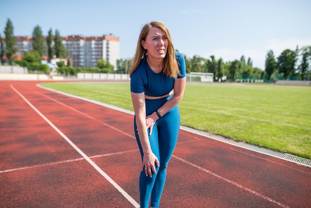 Mujer joven sosteniendo su pierna con dolor en el estadio Correr lesión deportiva fractura de la articulación dislocada Una mujer deportiva está tocando su pierna debido a un esguince