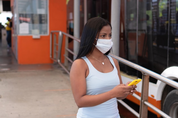 Mujer joven sosteniendo su teléfono mientras espera su autobús en la estación. Lleva una máscara debido a la pandemia de COVID-19.