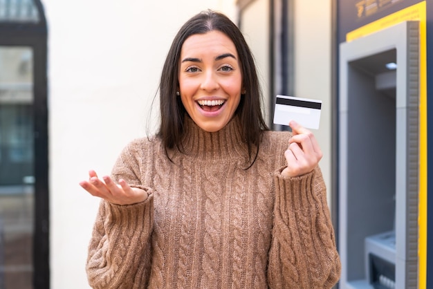 Mujer joven sosteniendo una tarjeta de crédito al aire libre con expresión facial sorprendida