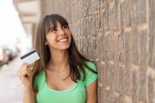 Foto mujer joven sosteniendo una tarjeta de crédito al aire libre mirando hacia arriba mientras sonríe