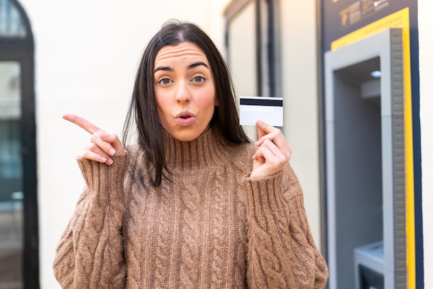 Mujer joven sosteniendo una tarjeta de crédito al aire libre sorprendida y apuntando hacia el lado
