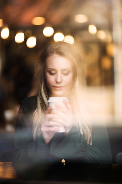 Mujer joven sosteniendo una taza de café en una cafetería vista a través del vidrio
