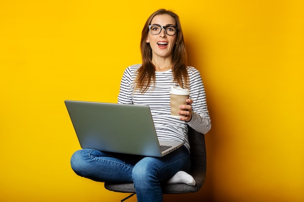 Mujer joven sosteniendo una taza de café, sentado en una silla, trabajando en una computadora portátil en un amarillo aislado.