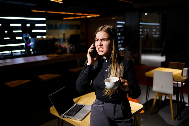 Mujer joven sosteniendo una taza de café y usando la computadora portátil hablando por teléfono. Empresaria trabajando desde casa. Trabajar desde casa