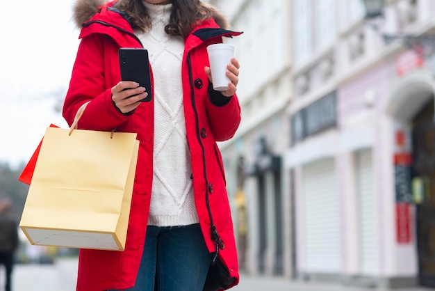 Mujer joven sosteniendo té y usando el teléfono al aire libre en la calle de invierno