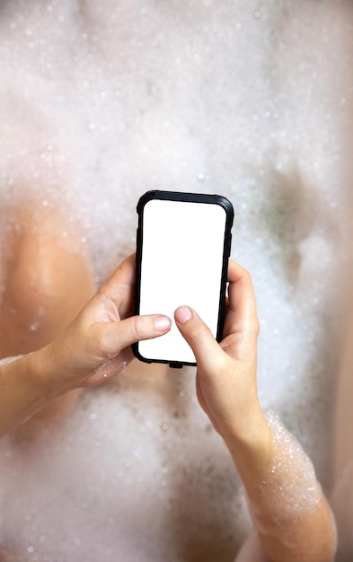 Mujer joven sosteniendo un teléfono inteligente en blanco en la bañera con fondo de vista superior de primer plano de espuma jabonosa
