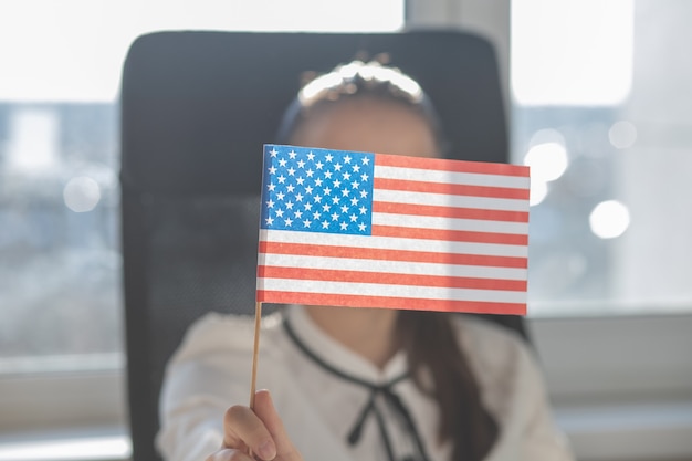 Mujer joven sostiene la bandera americana en su mano, se sienta en un sillón en una oficina en una mesa