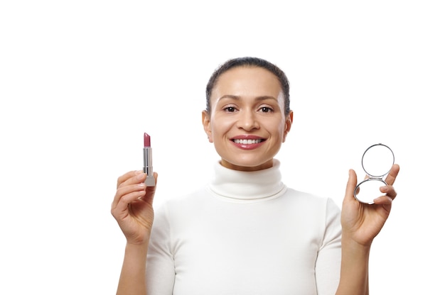 Mujer joven sostiene un lápiz labial y un espejo en las manos mientras posa con una hermosa sonrisa dentuda a la cámara