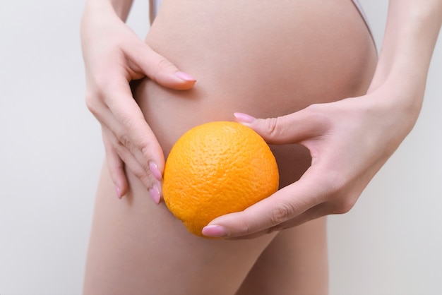 La mujer joven sostiene una naranja sobre un fondo claro. Concepto de problema de celulitis