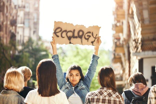 Mujer joven sostiene una pancarta con la palabra ecología mientras está de pie en el camino alrededor de activistas femeninas