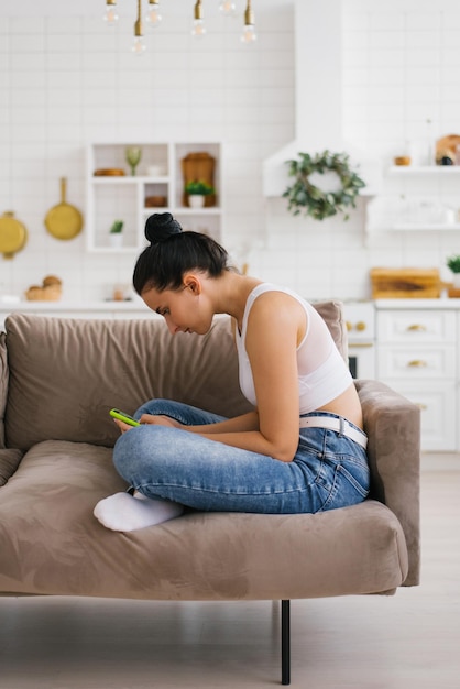 Mujer joven sostiene un teléfono inteligente en sus manos encorvada sentada en el sofá