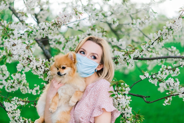 Mujer joven y spitz rojo con una máscara médica en su rostro en la naturaleza en un día de primavera. Pandemia de coronavirus