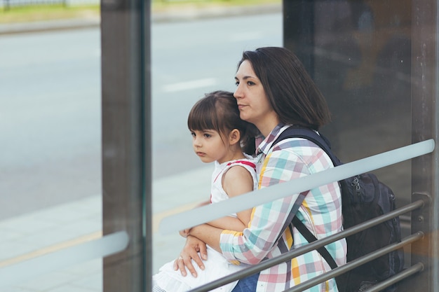 Una mujer joven con su hija está esperando un autobús público en la estación de autobuses