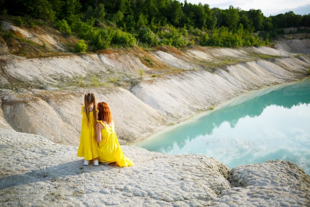 Mujer joven con su hija en un vestido amarillo cerca del lago con agua azul y árboles verdes. Concepto de relación familiar feliz