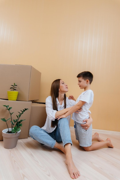 Una mujer joven con su hijo está sentada frente a los palcos y se regocija por la inauguración de la casa después de mudarse. Inauguración de la casa, entrega y transporte de carga, compra de bienes raíces.