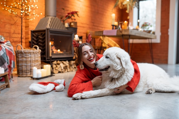 Mujer joven con su lindo perro blanco durante unas felices vacaciones de año nuevo sentado junto a una chimenea en casa