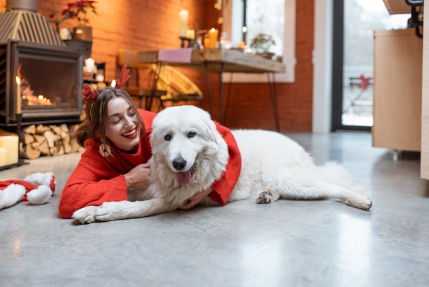 Mujer joven con su lindo perro blanco durante unas felices vacaciones de año nuevo sentado junto a una chimenea en casa