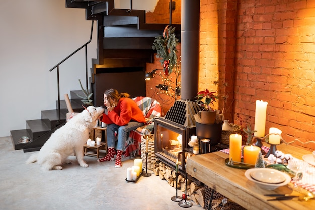 Mujer joven con su lindo perro blanco durante unas felices vacaciones de año nuevo sentado junto a una chimenea en casa