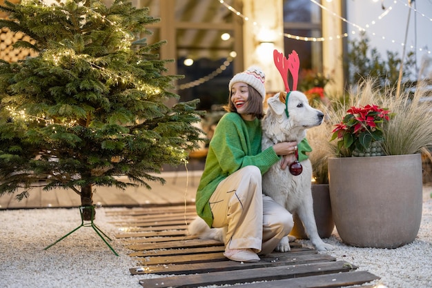 Mujer joven con su lindo perro blanco en el patio trasero decorado de la casa de campo de año nuevo. Chica divirtiéndose con un perro en las vacaciones de invierno. Perro con orejas de ciervo