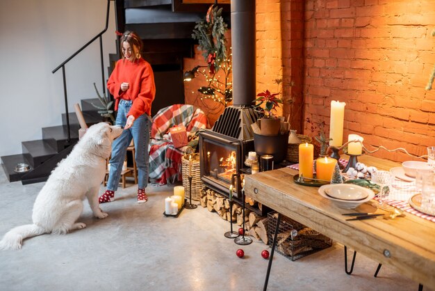 Mujer joven con su lindo perro preparándose para unas vacaciones de año nuevo junto a la chimenea y la mesa de comedor en casa