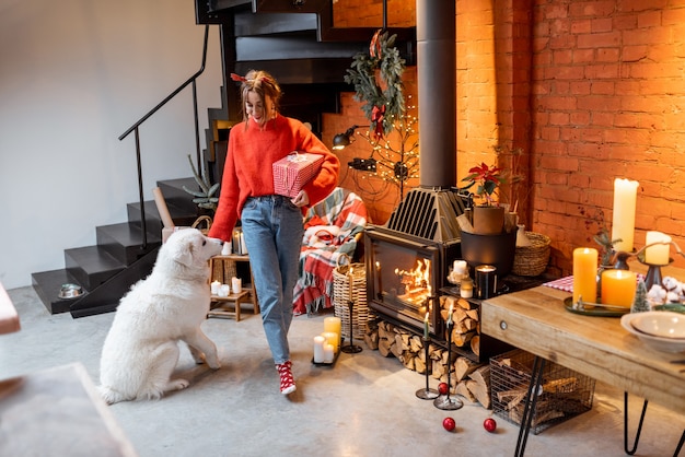 Mujer joven con su lindo perro preparándose para unas vacaciones de año nuevo junto a la chimenea y la mesa de comedor en casa