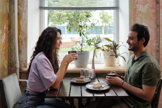 Mujer joven y su novio sonriente con tazas de café sentados junto a la mesa