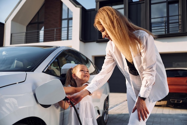 Mujer joven con su pequeña hija está con su coche eléctrico al aire libre