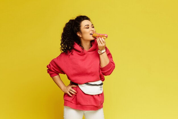 Foto mujer joven en una sudadera con capucha rosa comiendo un delicioso eclair sobre el fondo amarillo