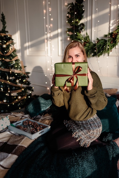 Una mujer joven con un suéter abre los regalos de Navidad en un dormitorio decorado para Navidad con el telón de fondo de un árbol de Navidad.