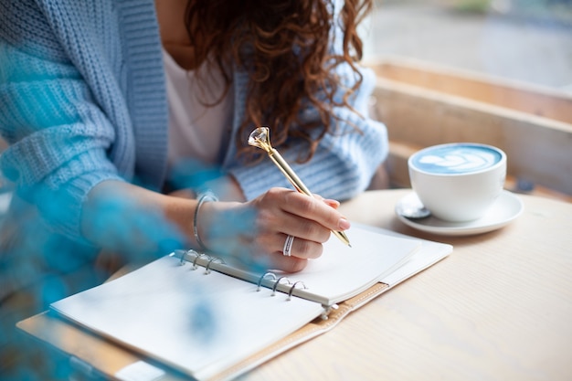 Mujer joven en suéter azul cálido sentado cerca del gran ventanal de la cafetería y escribiendo una lista de compras de Navidad con una taza de café con leche azul. Planificación de las vacaciones de Navidad. Concepto de organización y planificación.