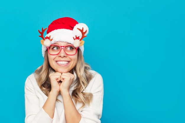 Una mujer joven con un suéter blanco, gorro de Papá Noel, gafas de Navidad, mirando a otro lado en el espacio vacío de la copia