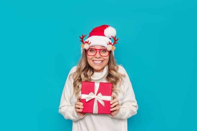 Una mujer joven en suéter blanco gorro de Papá Noel muestra una caja de regalo aislada sobre un fondo de color azul