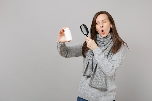 Mujer joven en suéter, bufanda mirando tabletas de medicamentos pastillas de aspirina en botella con lupa aislado sobre fondo gris. Estilo de vida saludable enfermedad enferma tratamiento concepto de estación fría.