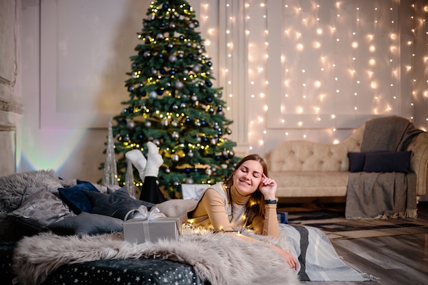 Una mujer joven con un suéter con un cabello largo y hermoso se sienta y se ríe frente a los regalos de Navidad. Feliz Año Nuevo